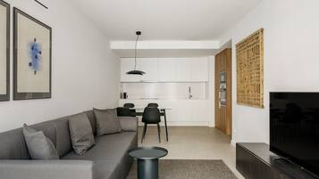 A modern living room with a grey sofa, a small round table, a flat-screen TV, and a kitchen in the background.