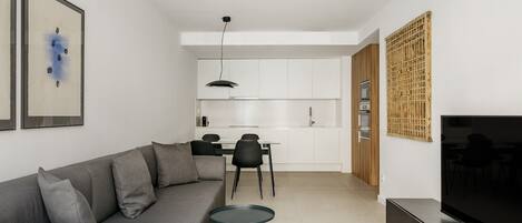 A modern living room with a grey sofa, a small round table, a flat-screen TV, and a kitchen in the background.