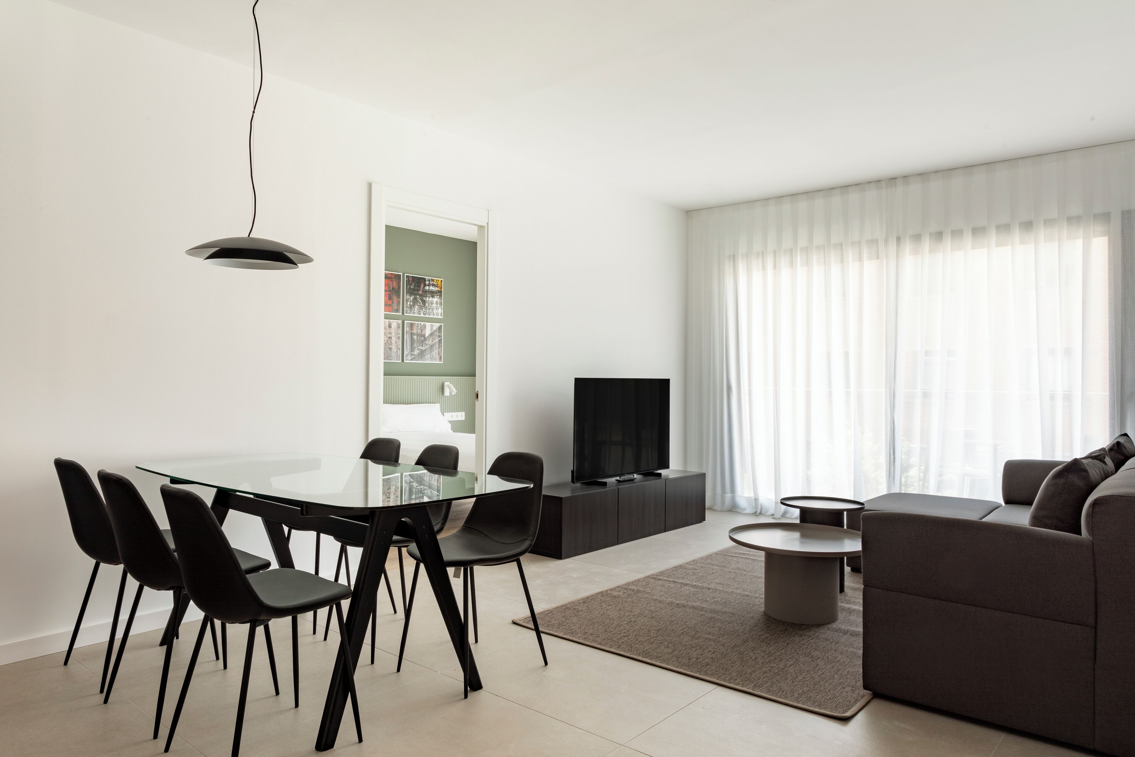 Un salon moderne avec une table à manger en verre, des chaises noires, un canapé gris, un téléviseur à écran plat et une grande fenêtre ornée de rideaux légers.
