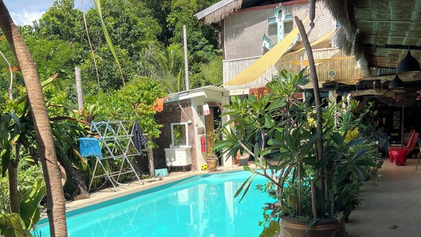 A pool area with a wooden lounge chair, potted plants, and a building with a thatched roof in the background.