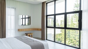 Una habitación de hotel moderna con una cama grande, una mesita de madera, un espejo y un ventanal con vistas a árboles.