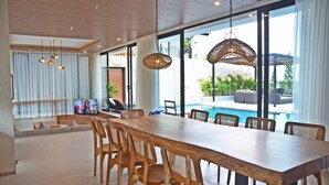 Un comedor amplio con una larga mesa de madera y sillas, ventanas grandes que ofrecen vista a una piscina y follaje exterior.