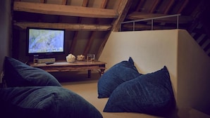 Een knus zolderkamer met een houten bureau, een televisie en blauwe kussens.