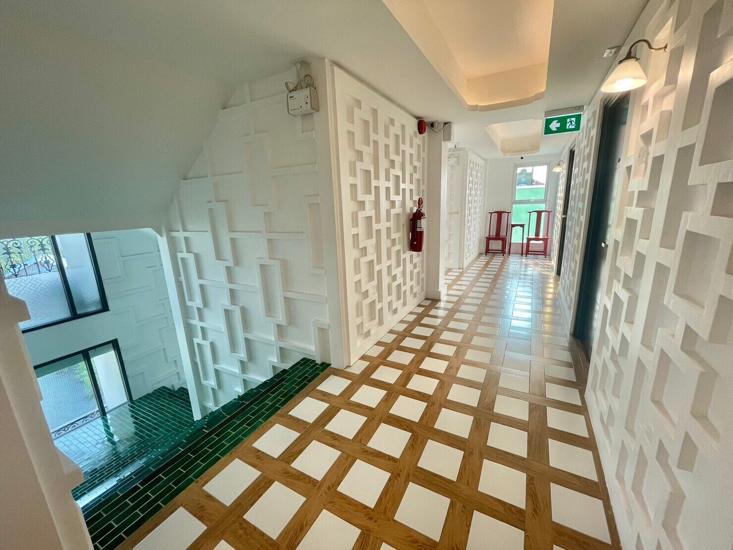 A hallway with patterned walls and a checkered floor, leading to a staircase and a door with a green exit sign.