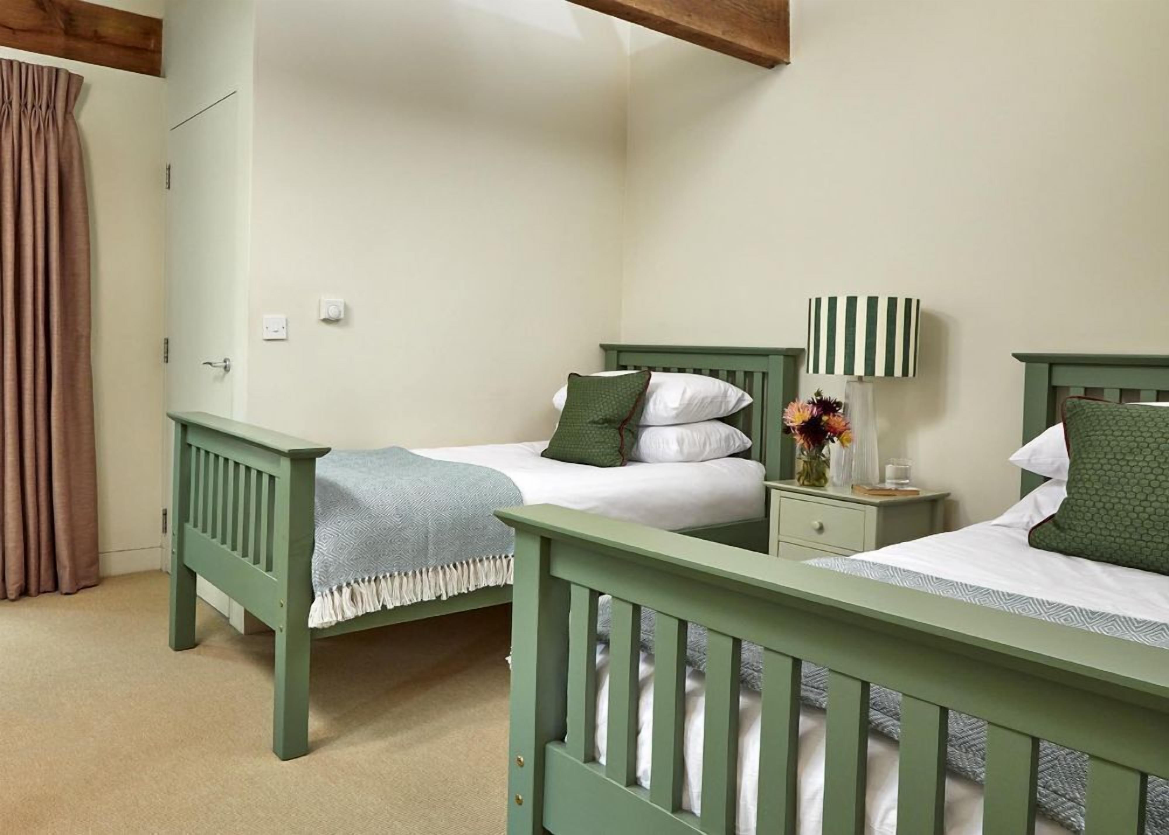 Un dormitorio con dos camas de madera verde, ropa de cama blanca, una mesita de noche con una lámpara y un florero con flores.