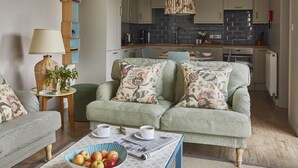 A cozy living room with a sofa and armchairs, a coffee table with apples and a newspaper, and a kitchen in the background.