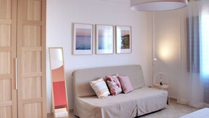 A bedroom with a beige sofa, three framed pictures on the wall, a bedside table with a lamp, and a wooden wardrobe.