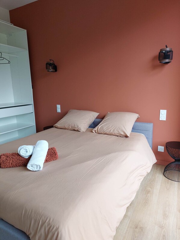 A neatly made bed with beige bedding, two pillows, and rolled towels. There is a small round table with a wireframe design next to the bed.