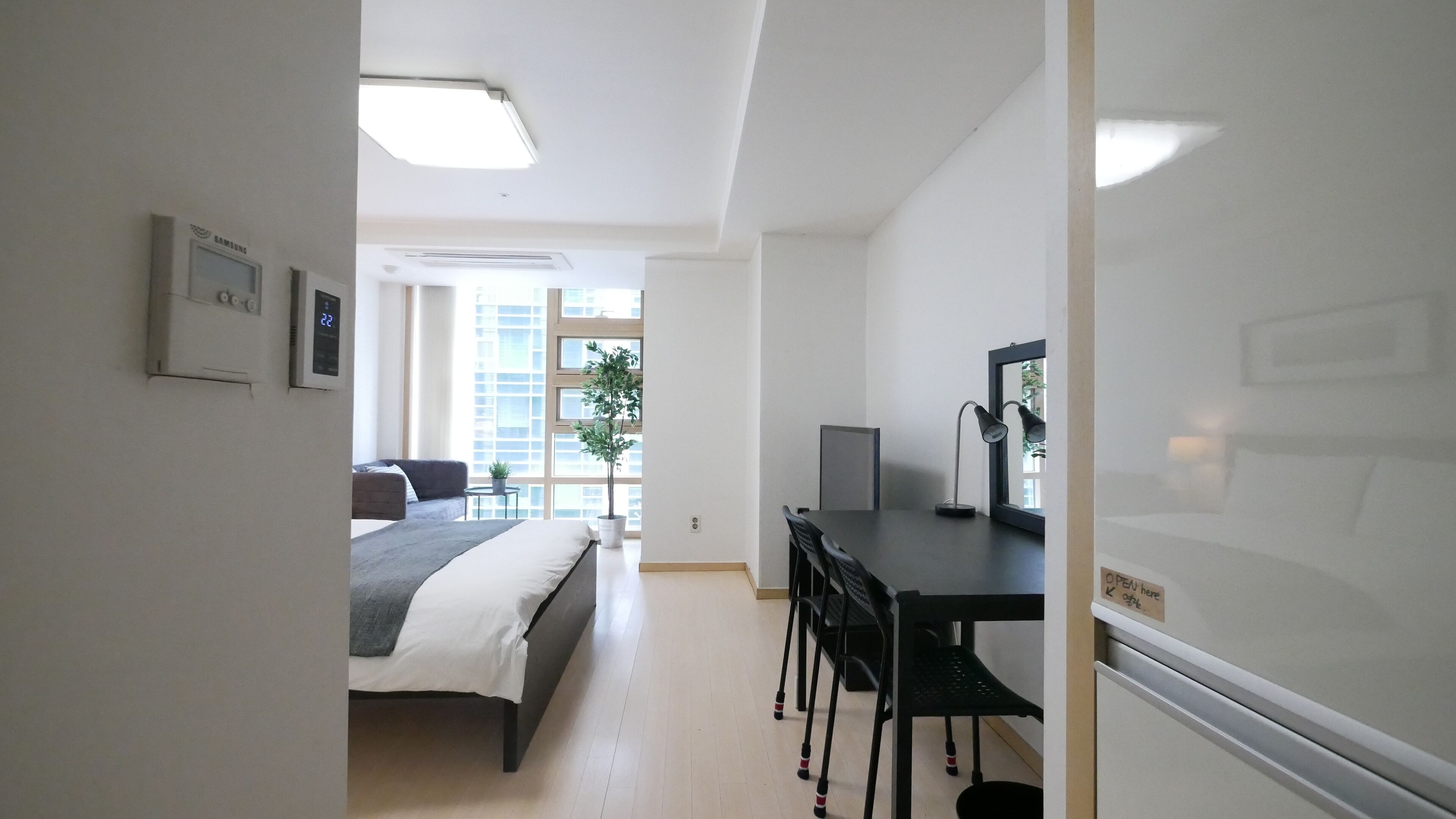 Une chambre d’hôtel moderne, équipée d’un lit, d’un bureau et de chaises. Une fenêtre offre une vue sur les bâtiments, et une plante est posée sur le rebord de la fenêtre.