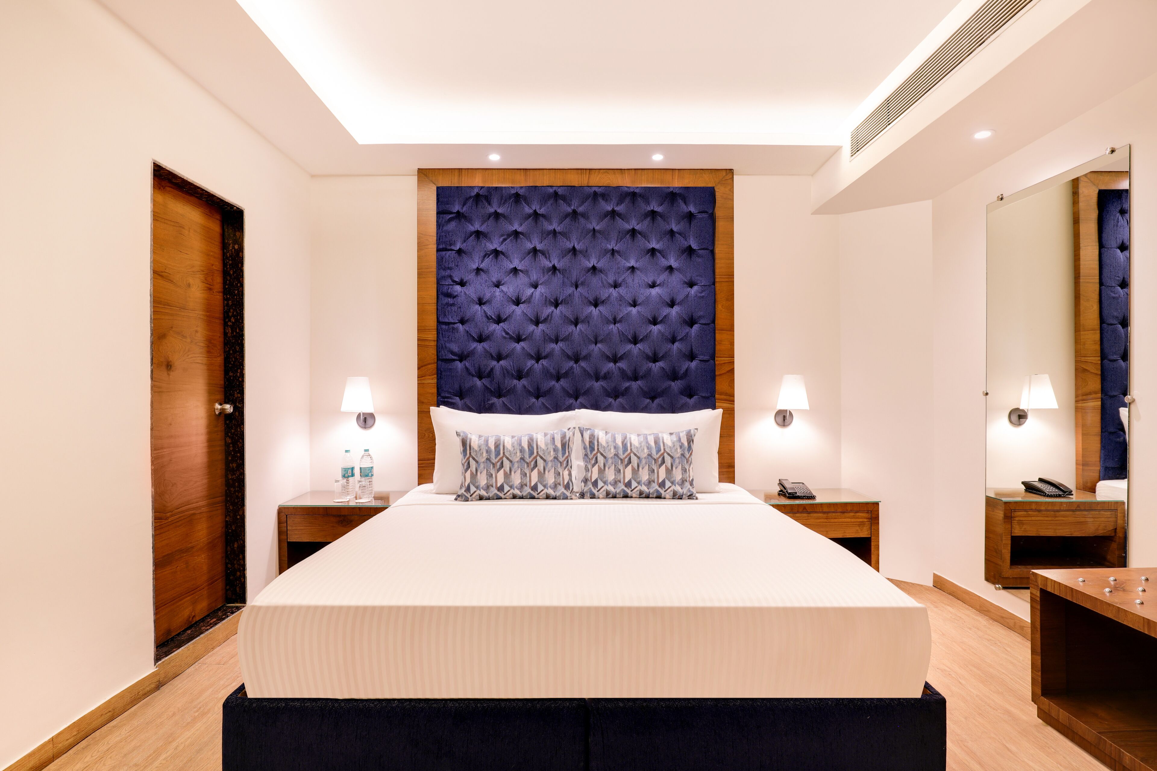 Habitación de hotel con una cama grande, dos mesitas de noche, una puerta de madera, un espejo y una rejilla de ventilación en el techo.