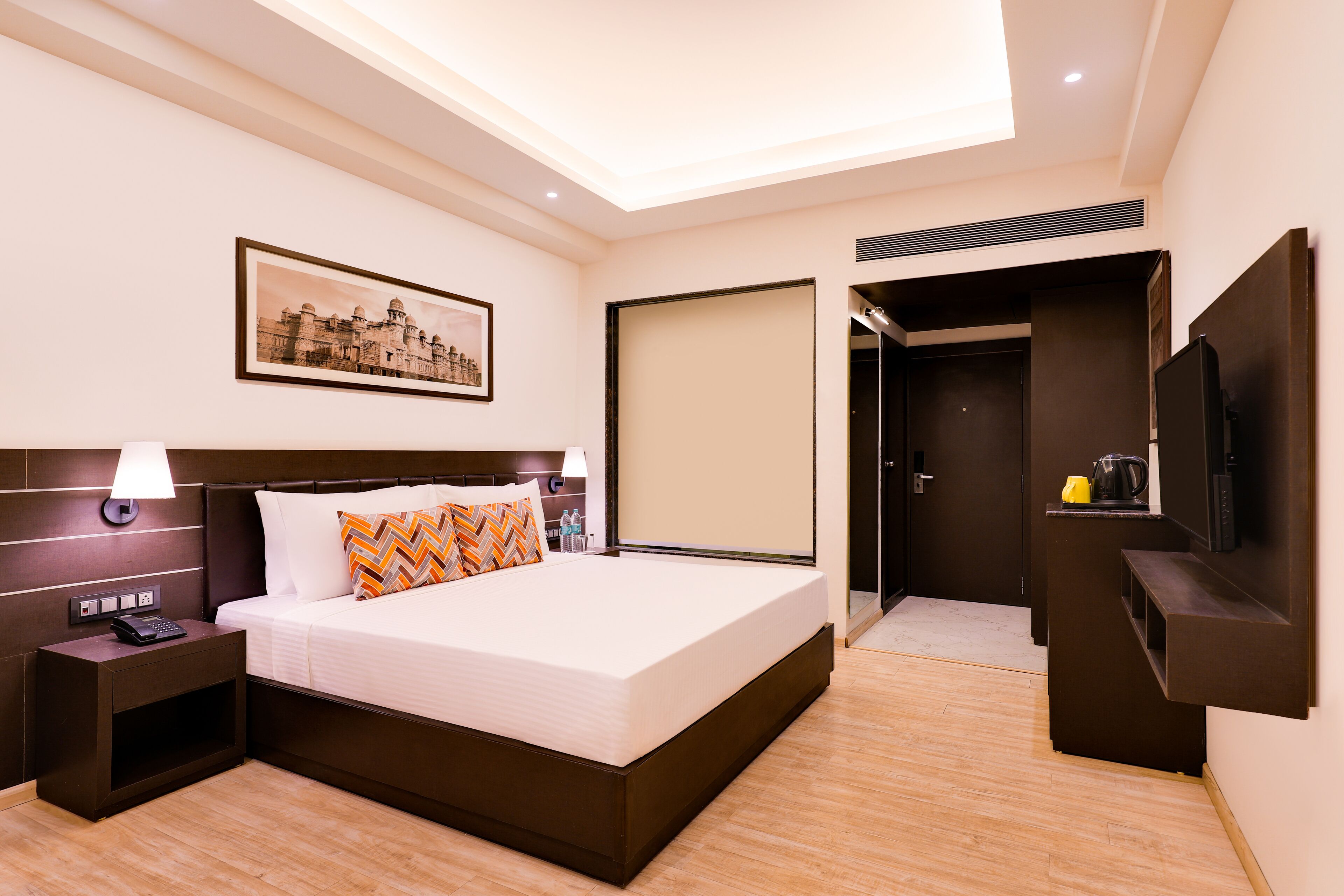 A modern hotel room with a large bed, a bedside table with a lamp, a flat-screen TV, and a painting on the wall.