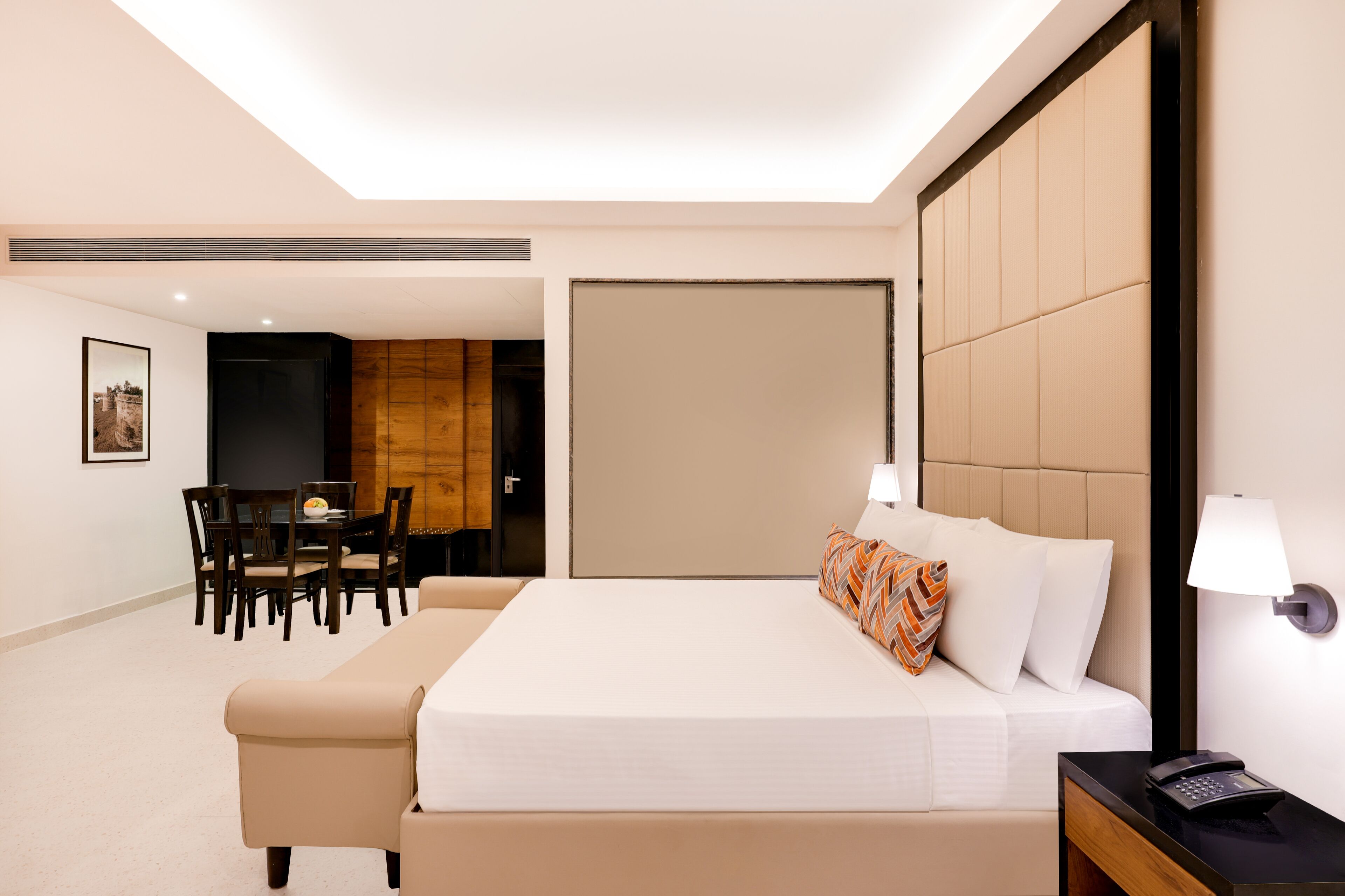 A hotel room with a large bed, a bedside table with a lamp, a dining area with a table and chairs, and a wooden panel wall.