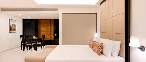 A hotel room with a large bed, a bedside table with a lamp, a dining area with a table and chairs, and a wooden panel wall.