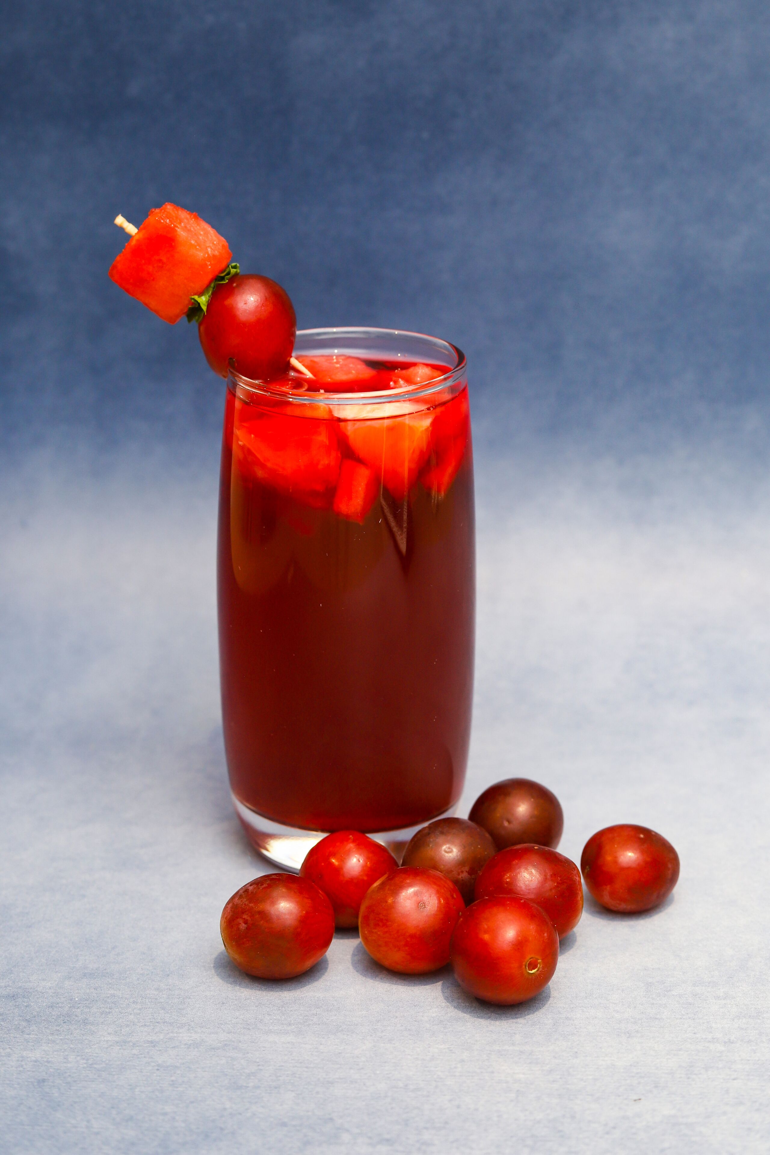 A tall glass with a red beverage, garnished with a cherry and a slice of watermelon.
