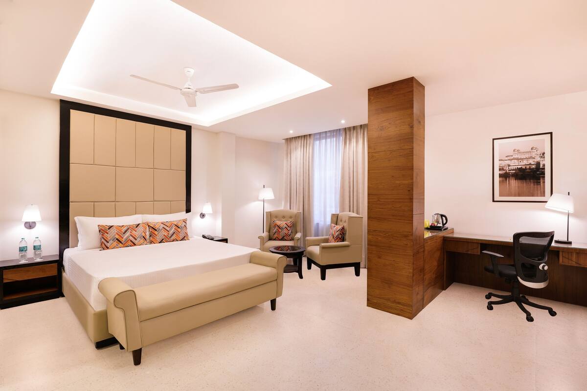 Habitación de hotel con una cama grande, un escritorio, dos sillones, un ventilador de techo y vistas a un edificio a través de la ventana.