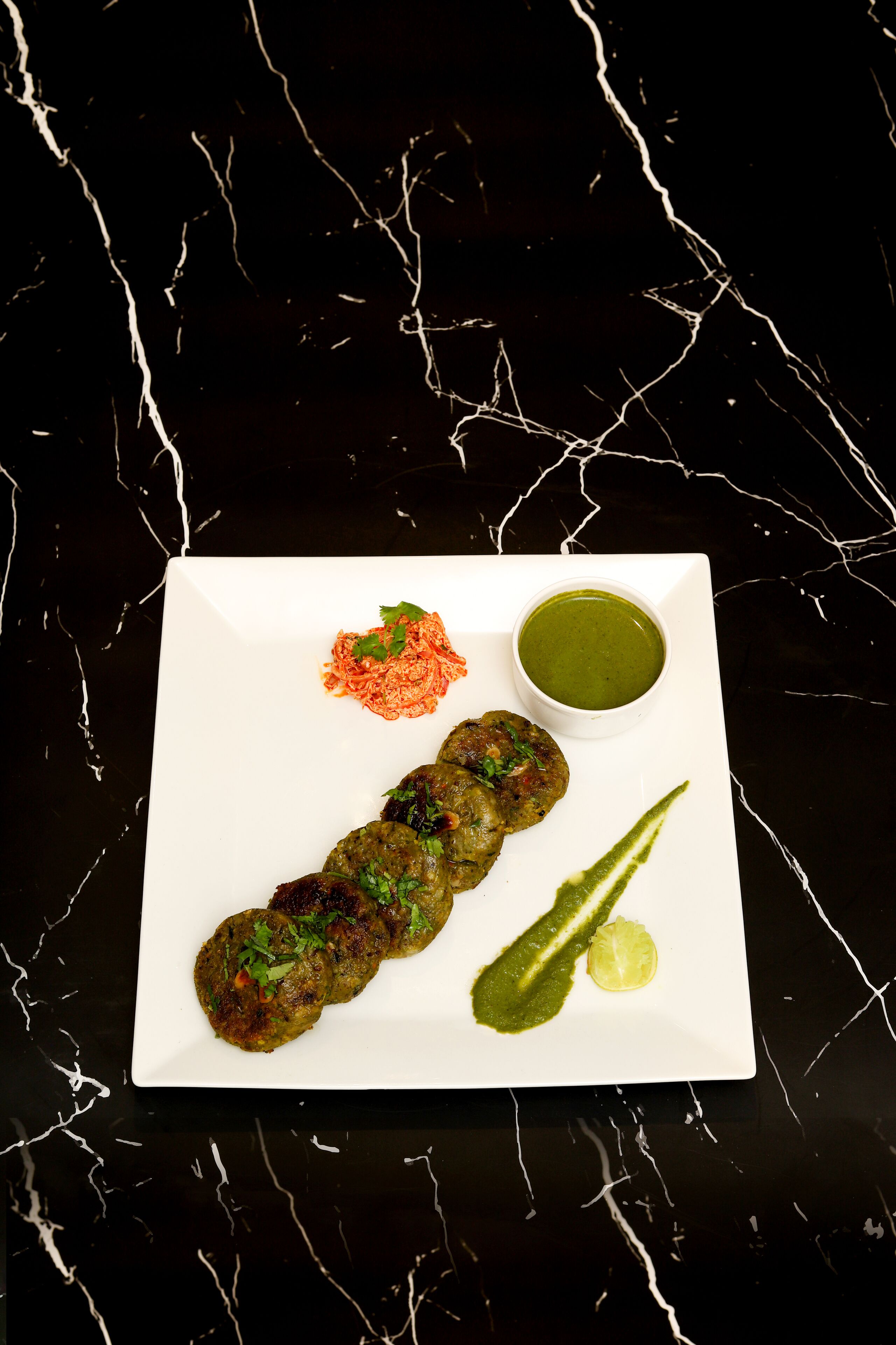 A square plate with three meat patties, a small bowl of green sauce, and a wedge of lime on a black marble surface.