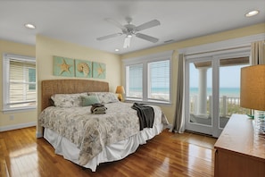 A bedroom with a large bed, a ceiling fan, and a view of the ocean through a sliding glass door.