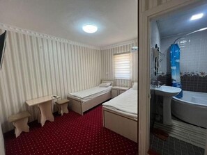 Habitación de hotel compacta con dos camas, un escritorio pequeño, una silla y un baño con lavamanos y bañera.