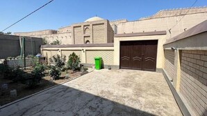 A courtyard with a large building featuring a dome and arched windows, a gated entrance, and a green trash bin. - Shodlik Guest House (Khiva)