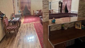 A spacious living area with a wooden floor, a sofa, a dining table with chairs, and a red carpet. There is a large mirror on the wall and a clock on the wall. - Shodlik Guest House (Khiva)