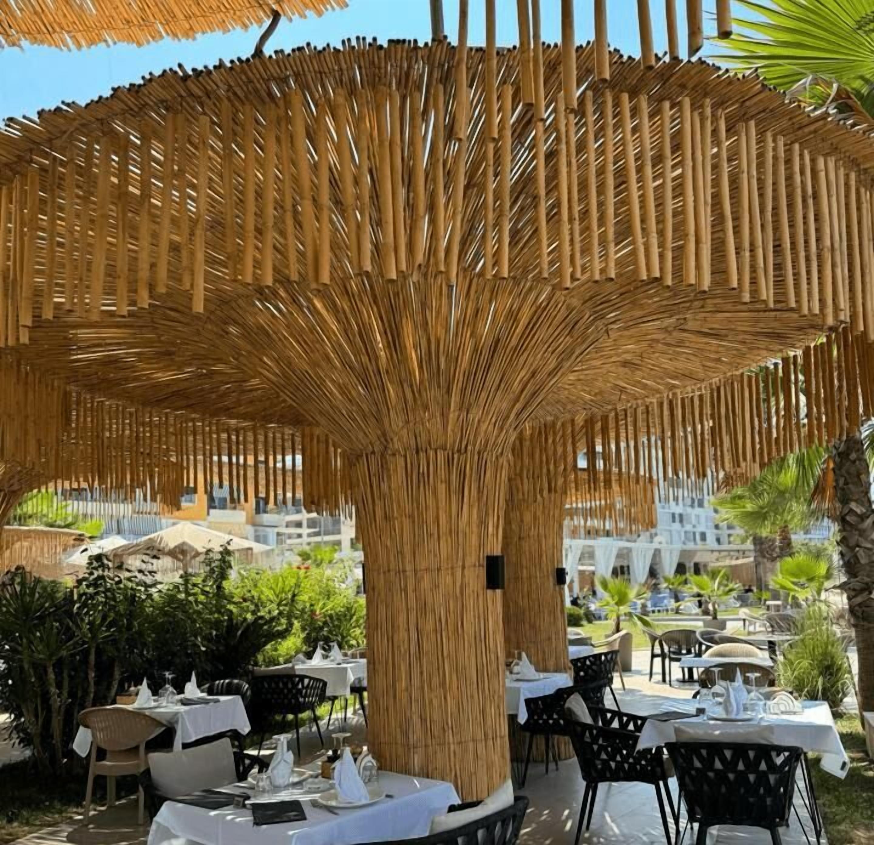 Un área de comedor al aire libre con estructuras de bambú en el techo, mesas con manteles blancos y sillas negras.