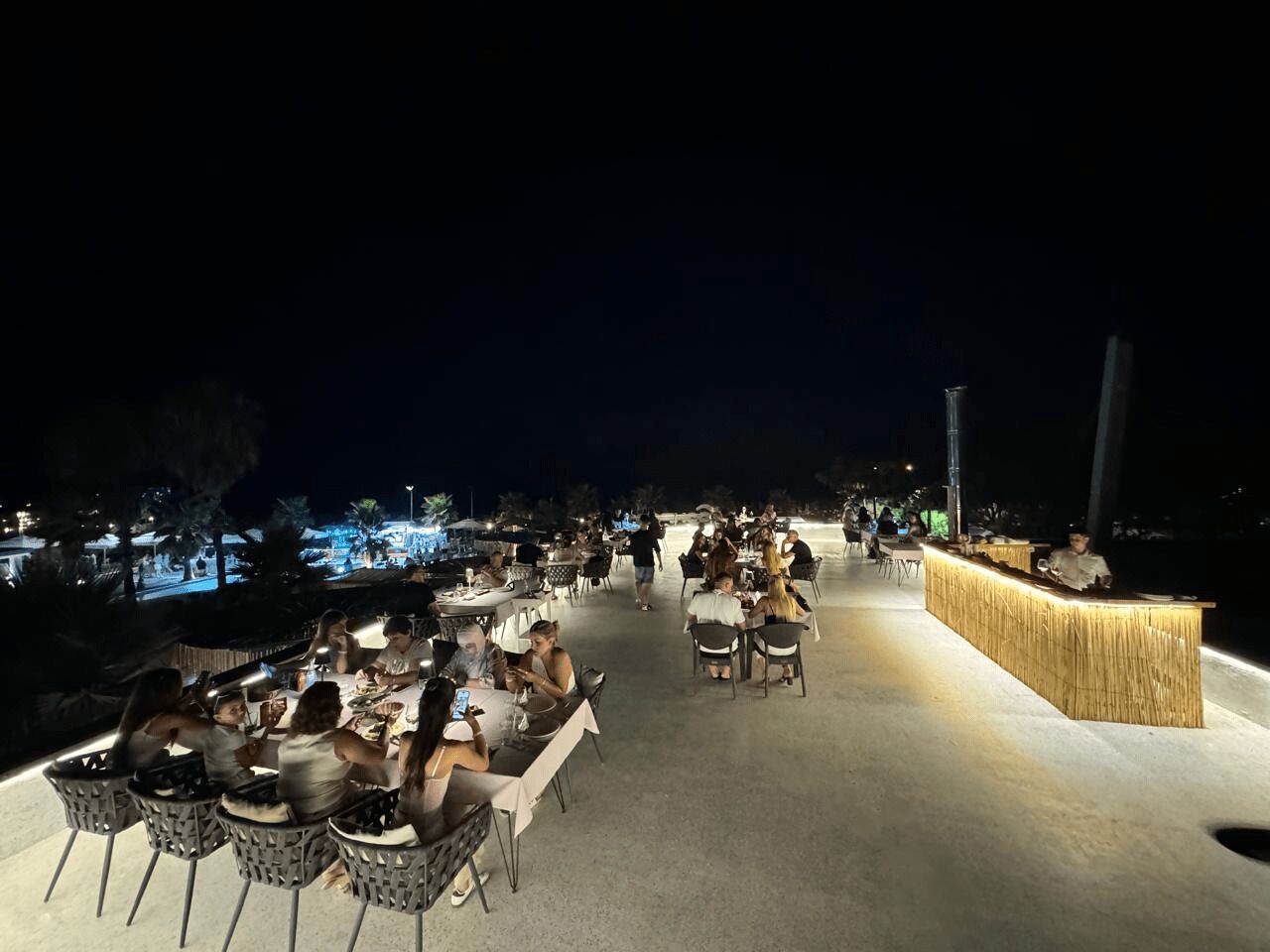 En udendørs spiseplads med borde og stole, en bar samt udsigt til stranden om natten.