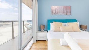 Quarto com uma janela grande, cama com roupa de cama branca, cabeceira azul e um quadro na parede.