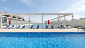 Uma área de piscina com espreguiçadeiras azuis e guarda-sóis vermelhos, com vista para um corpo de água e um navio de cruzeiro.