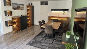 Un espace repas moderne avec une table en bois, des chaises foncées et un tapis noir. Des plantes en pot sont disposées sur le côté, et un coin cuisine intégré comprend un meuble et des étagères en bois.