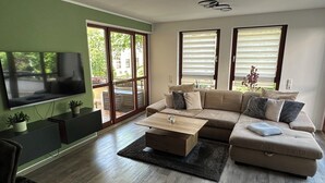 Un salon moderne avec un canapé d’angle, un téléviseur à écran plat et de grandes fenêtres.