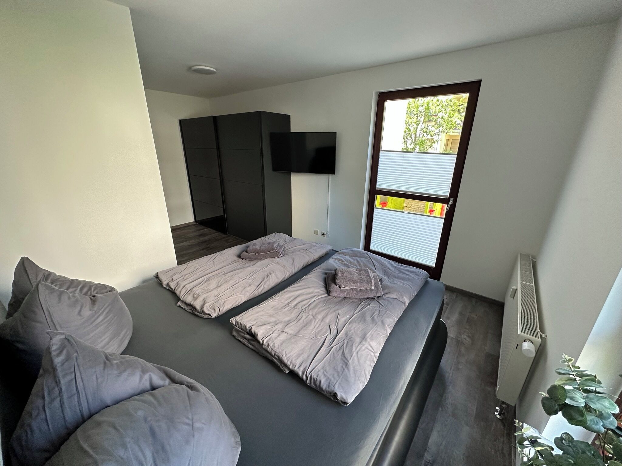 A modern bedroom with a large bed, a TV mounted on the wall, and a window with blinds.