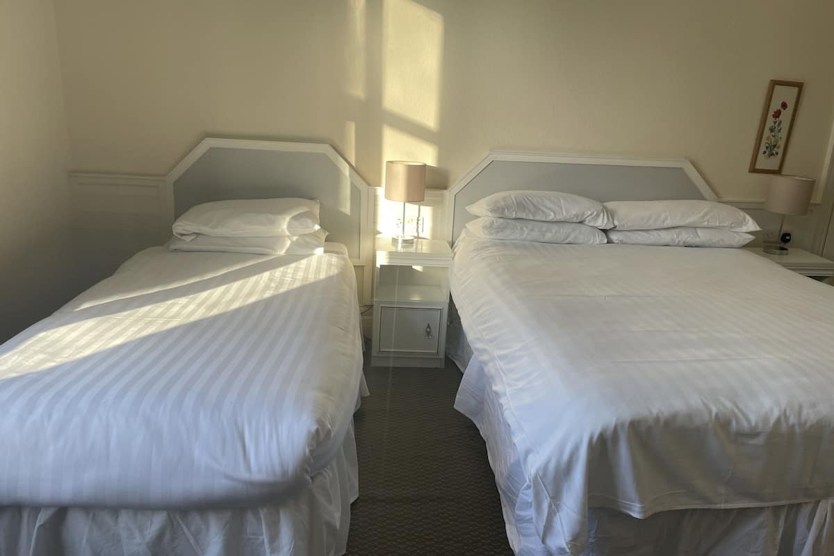 Ein Hotelzimmer mit zwei Einzelbetten, jeweils mit weißem Kopfteil und passenden Kissen. Dazwischen befindet sich ein Nachttisch mit einer Lampe, und an der rechten Wand hängt ein gerahmtes Bild.