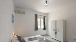 Un dormitorio con cama, armario, ventana con cortinas, lámpara de noche y aire acondicionado.