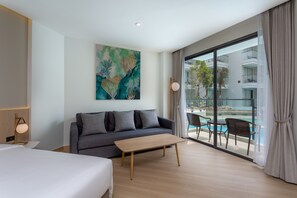Ein modernes Hotelzimmer mit einem großen Bett, einem grauen Sofa, einem Holztisch und Blick auf einen Swimmingpool durch raumhohe Fenster.
