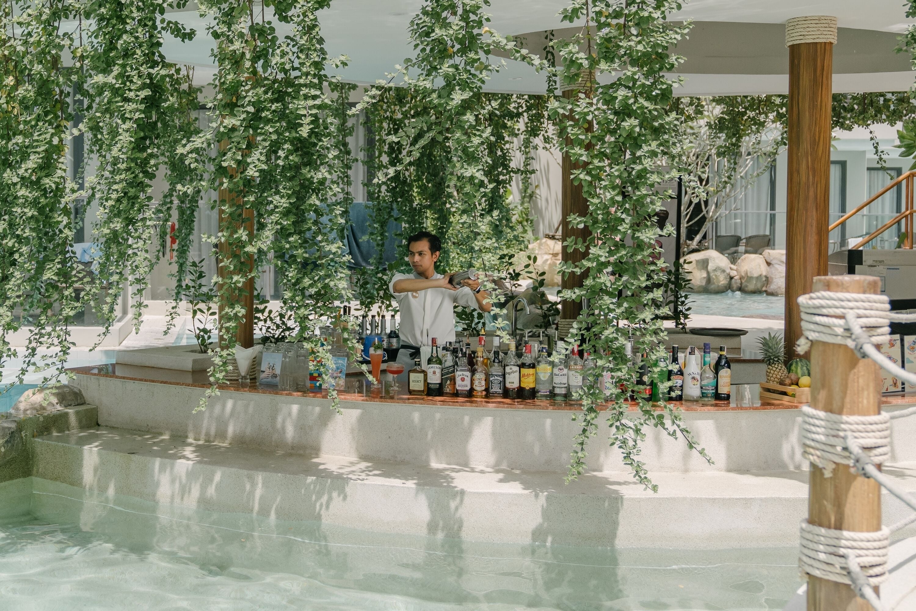 Zona de bar interior con piscina, rodeada de plantas colgantes y un pilar de madera.
