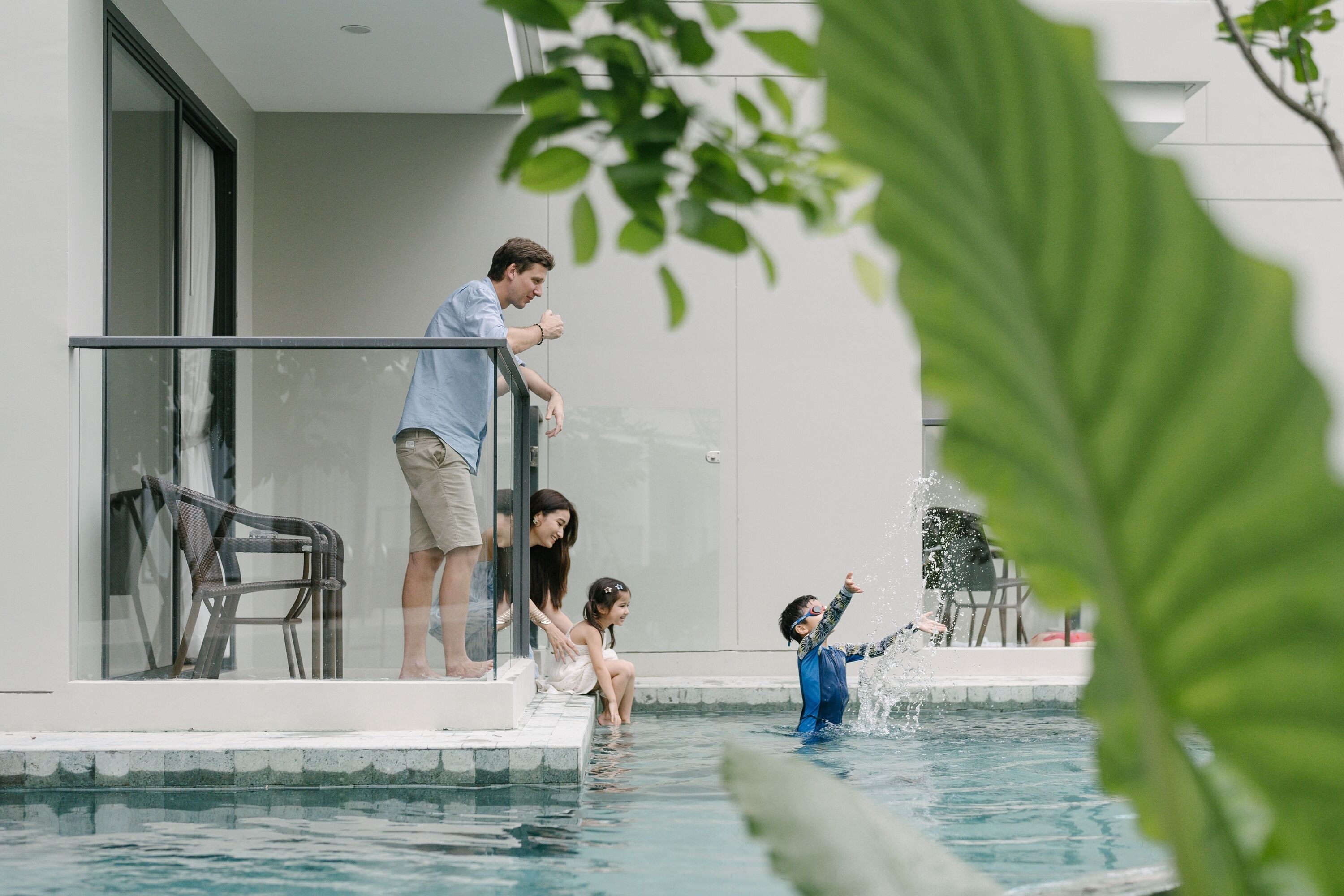 A modern pool area with a glass balustrade, a lounge chair, and a child playing in the water.