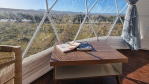 A geodesic dome tent with a table and a chair inside, overlooking a landscape with sparse vegetation and distant mountains.