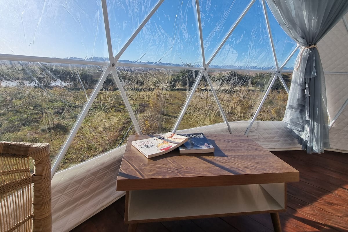 Una tienda en forma de domo geodésico con una mesa y una silla en su interior, con vistas a un paisaje de vegetación escasa y montañas distantes.