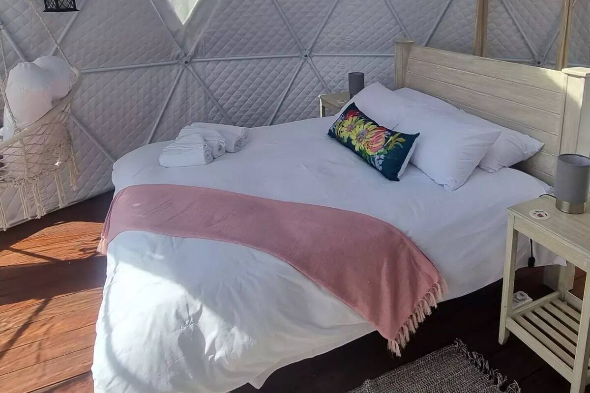 Una habitación tipo tienda acogedora y bien iluminada, con una cama bien hecha, una mesita de noche pequeña y una ventana que deja entrar luz natural.
