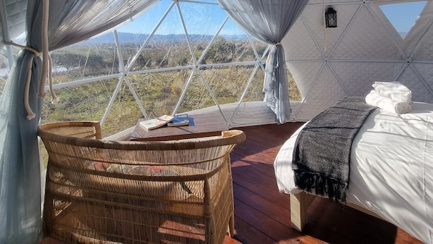 A geodesic dome tent with a bed, a wicker chair, and a view of the landscape.