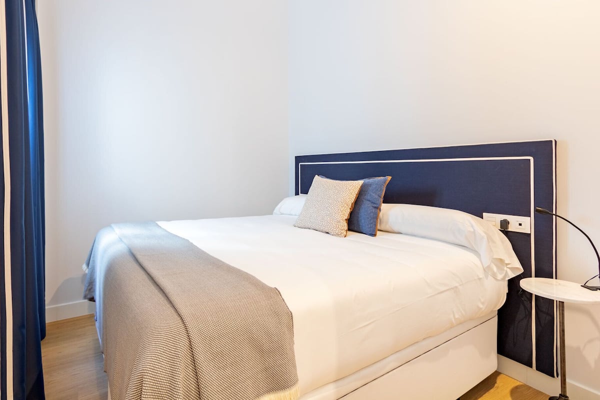 A neatly made bed with a white comforter and two pillows, a bedside table with a lamp, and a dark headboard.