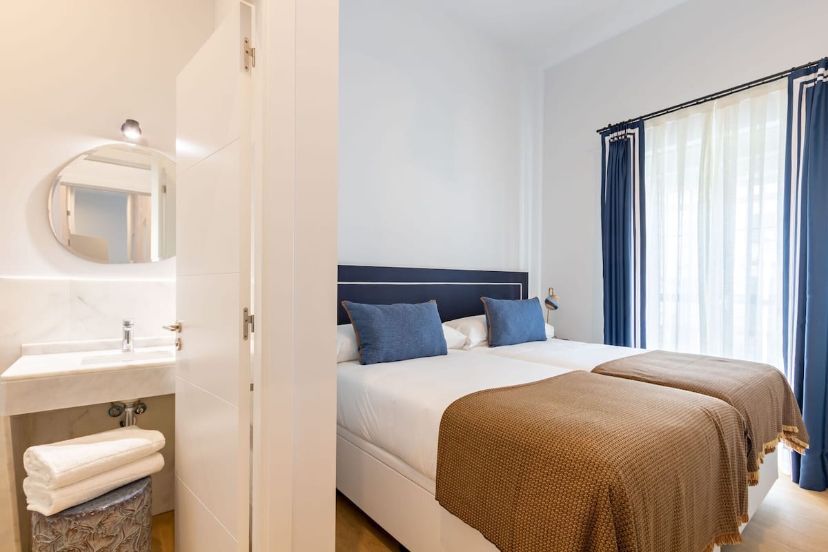 A hotel room with a white vanity, a round mirror, a bed with blue and brown bedding, and a window with blue curtains.