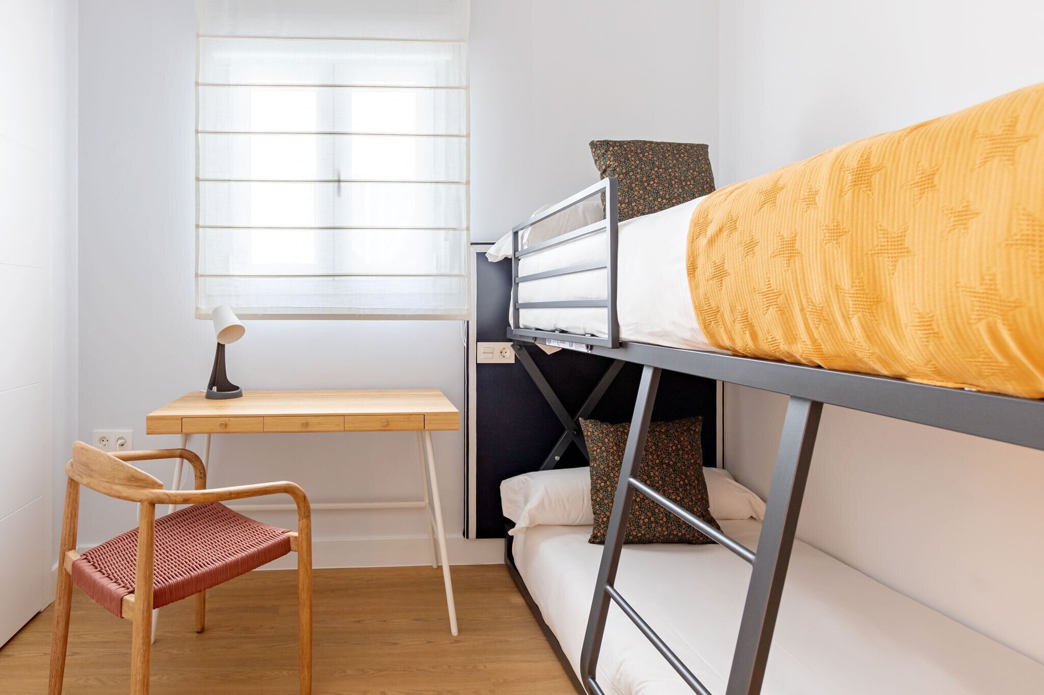 A bunk bed with a yellow patterned blanket, a desk with a lamp, and a chair.