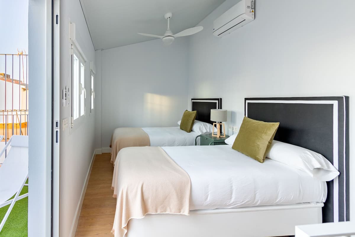 A hotel room with two beds, a ceiling fan, an air conditioning unit, and a window with a view of the outside.