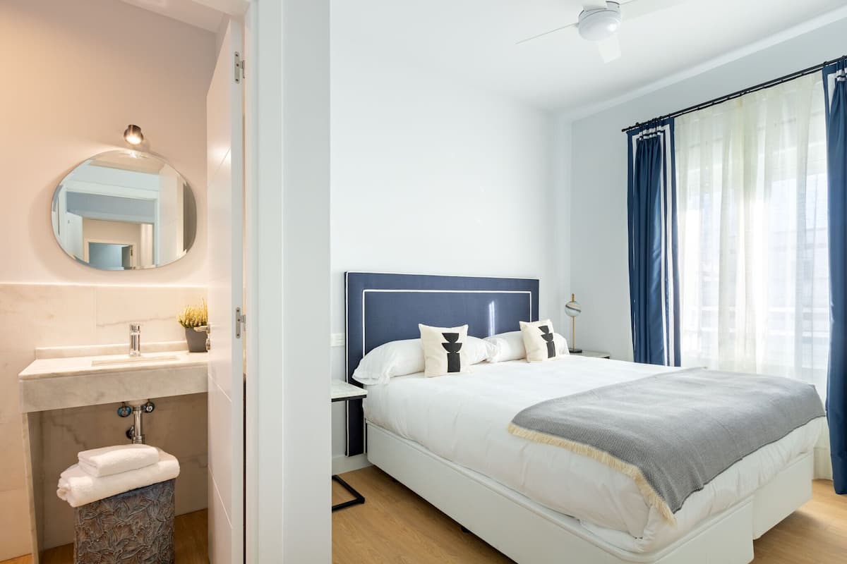 A modern bedroom with a large bed, a round mirror, a sink, and a window with curtains.