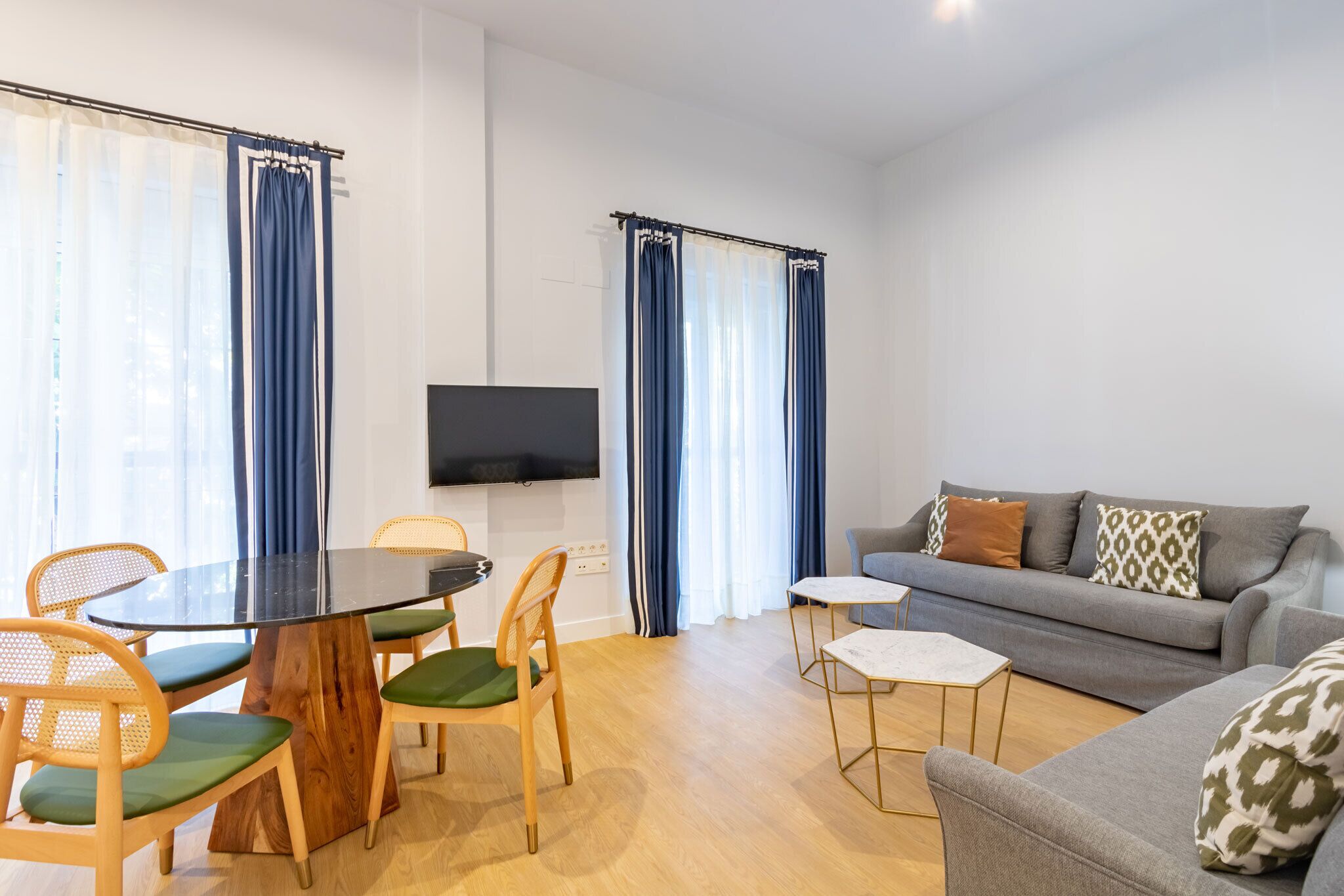 Una sala de estar moderna con una mesa de comedor redonda, sillas de madera, un sofá gris y un televisor de pantalla plana montado en la pared.
