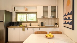 A modern kitchen with white cabinets, a stainless steel refrigerator, a microwave, and a dining table with chairs. There is a window providing natural light and a framed artwork on the wall. - MAMAD - Pet-friendly 2BR Apt & Kitchen (Tel Aviv)