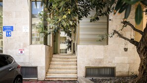 A building entrance with a staircase leading to a door, a car parked outside, and a tree with hanging pots. - MAMAD - Pet-friendly 2BR Apt & Kitchen (Tel Aviv)