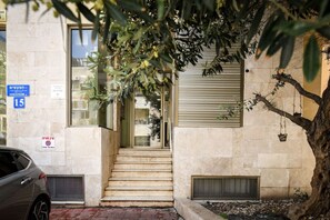 A building entrance with a staircase leading to a door, a parked car, and a tree in the foreground. - MAMAD - Pet-friendly 2BR Apt & Kitchen (Tel Aviv)
