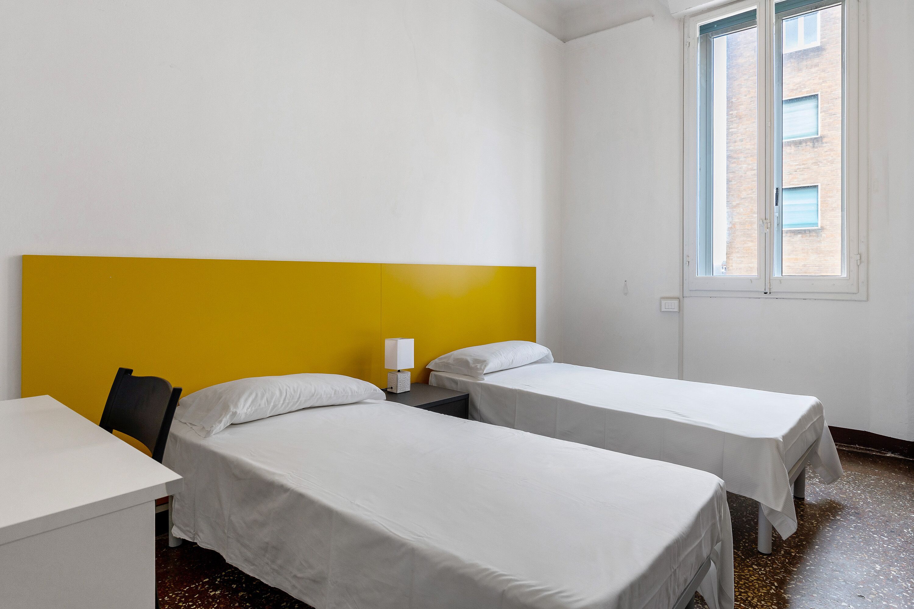 Een hotelkamer met twee enkele bedden, een bureau, een stoel en een raam met uitzicht op gebouwen.